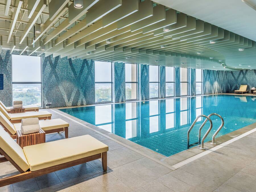 An indoor swimming pool with clear blue water, sun loungers, and a view of the city through large windows.