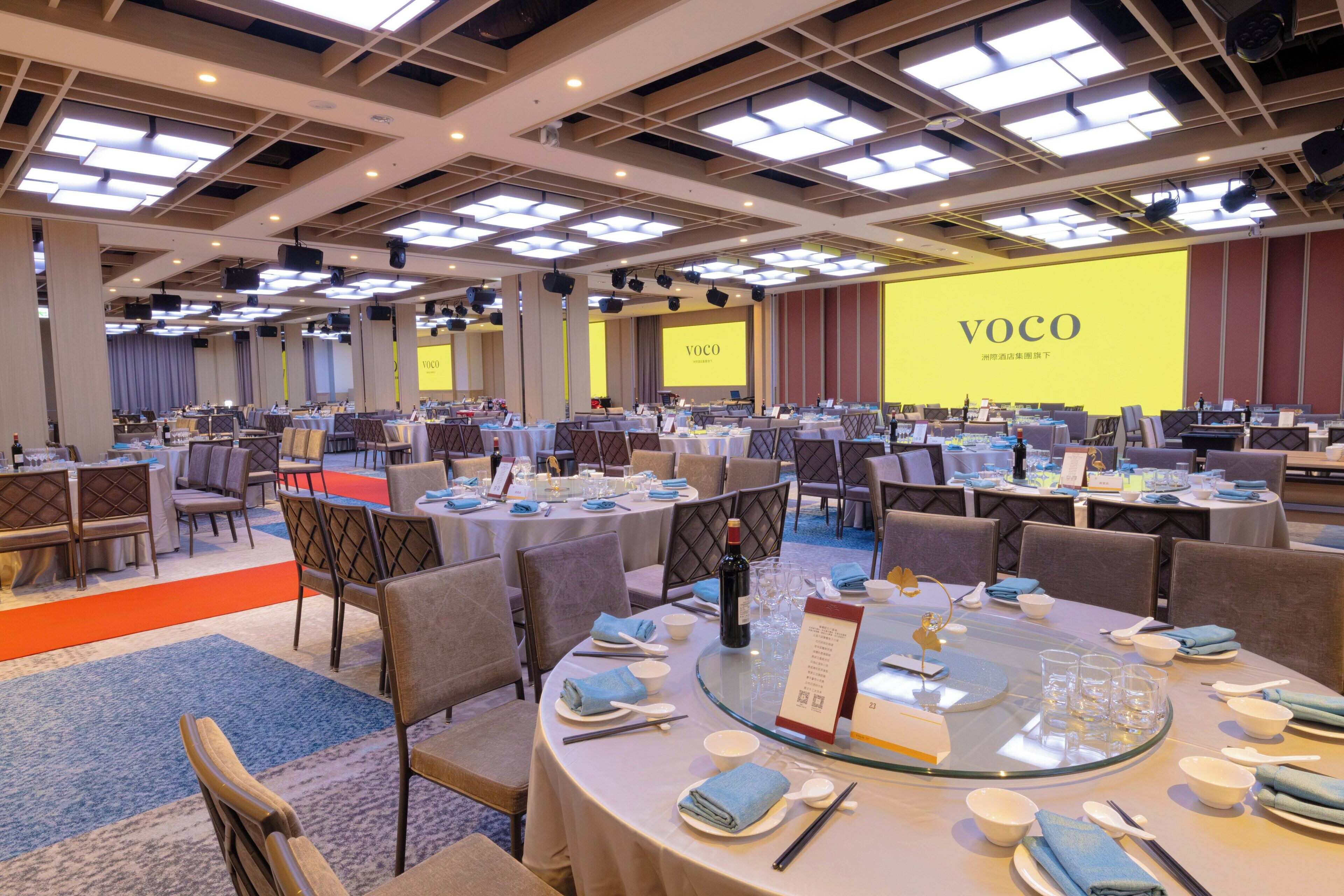 A conference room with multiple tables set for a meeting, equipped with microphones and a large screen displaying the word 'VOCO'.