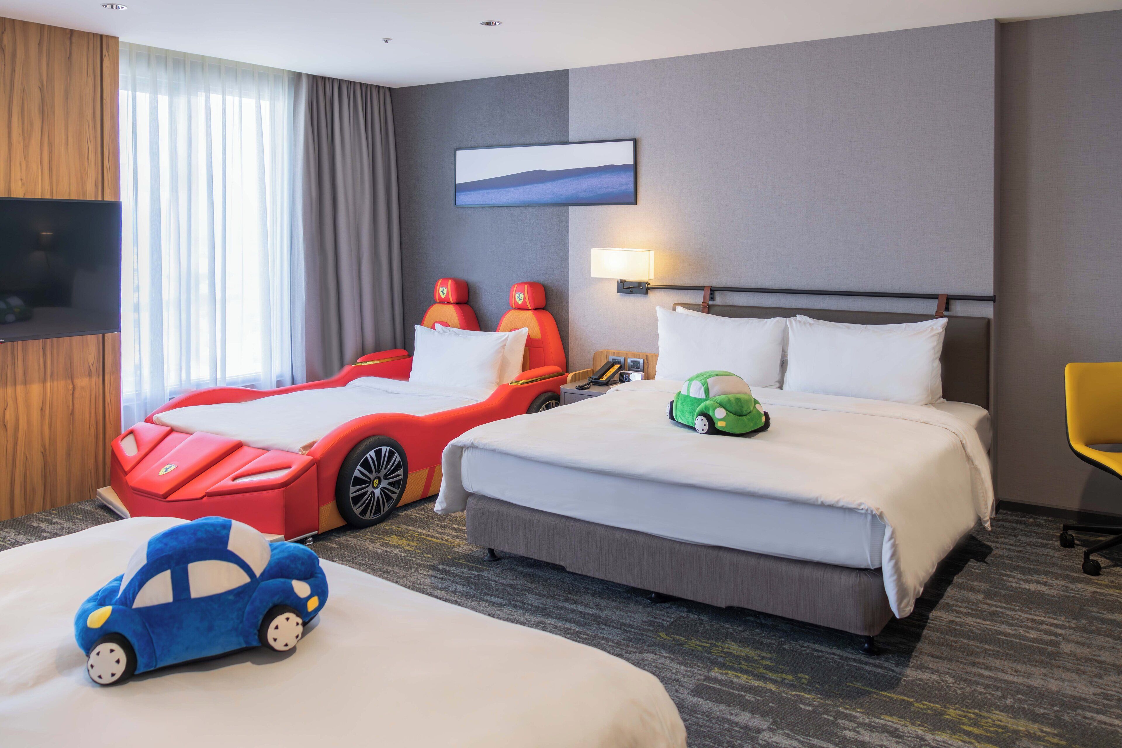 Ein Hotelzimmer mit einem großen Bett, einem kleineren Bett, einem Fernseher, einem Schreibtisch und einem Fenster mit Vorhängen.