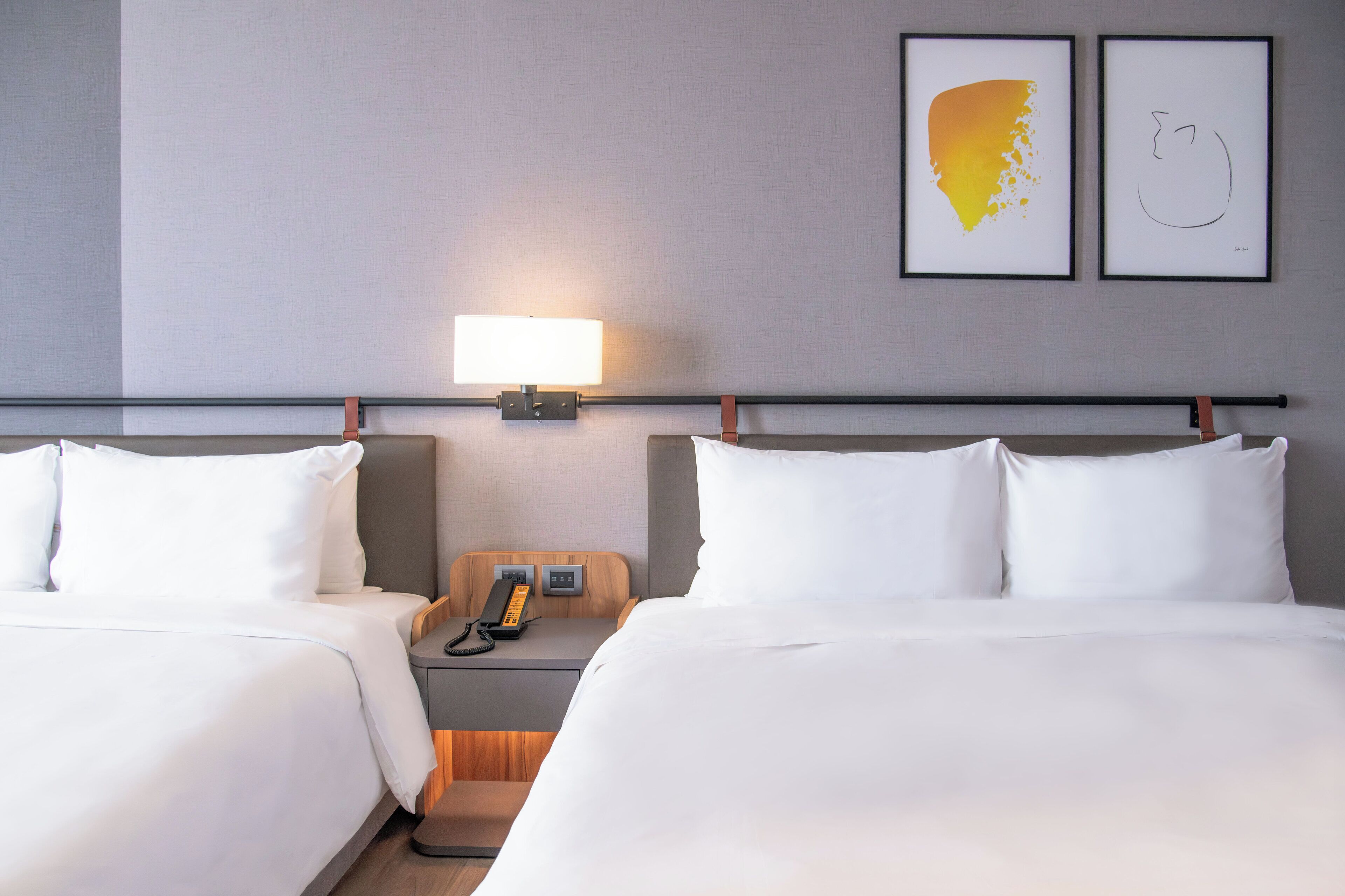 Hotellihuone, jossa on kaksi sänkyä, yöpöytä lampun kanssa, puhelin ja taideteos seinällä.