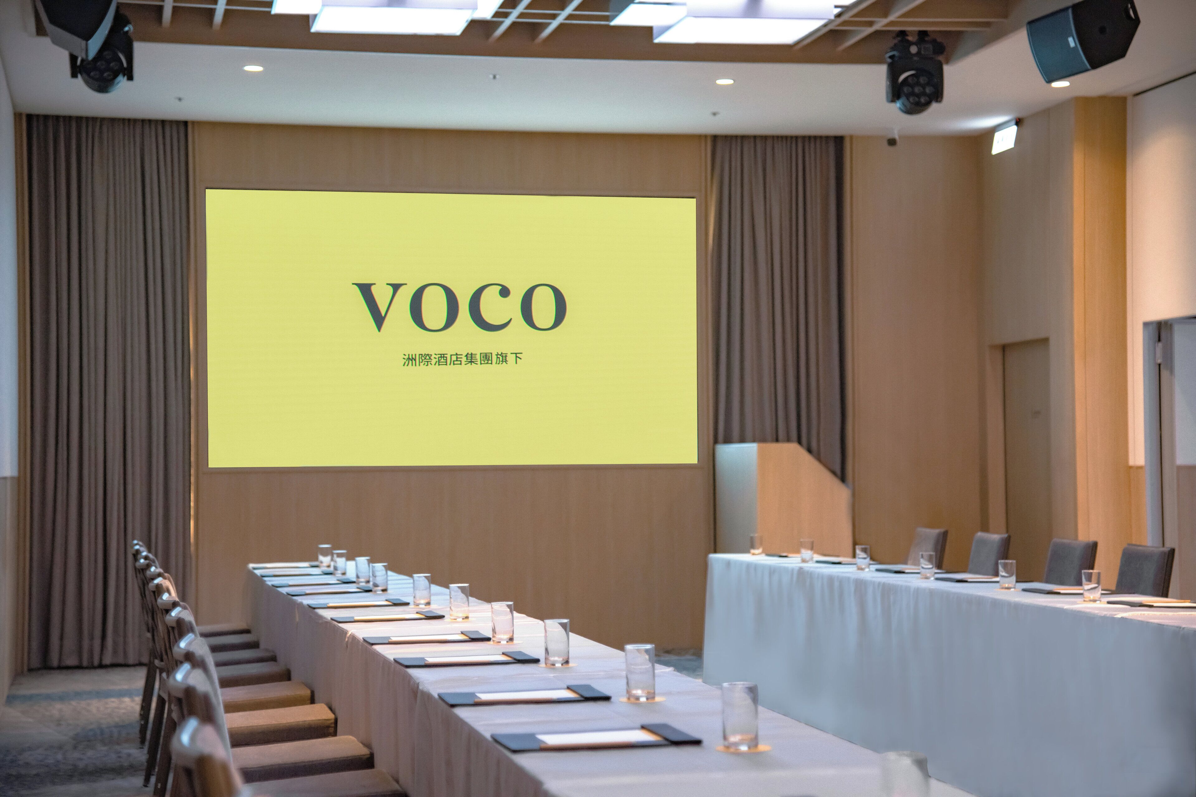 Un salón de conferencias con una larga mesa dispuesta para reuniones, una pantalla grande mostrando la palabra 'VOCO' y un sistema de sonido en el techo.