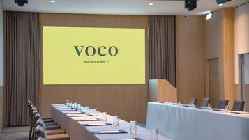 Un salón de conferencias con una larga mesa dispuesta para reuniones, una pantalla grande mostrando la palabra 'VOCO' y un sistema de sonido en el techo.