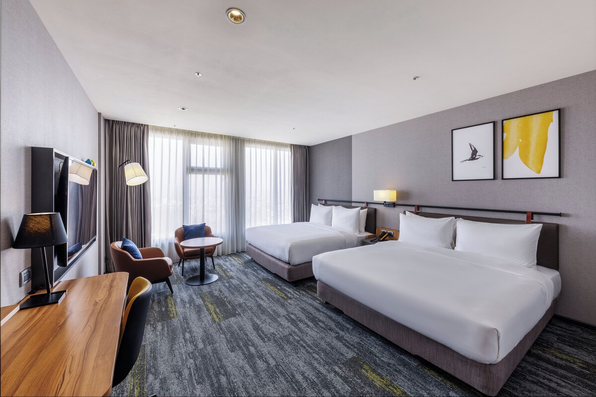 A hotel room with two beds, a desk, a chair, a TV, and a window with curtains.