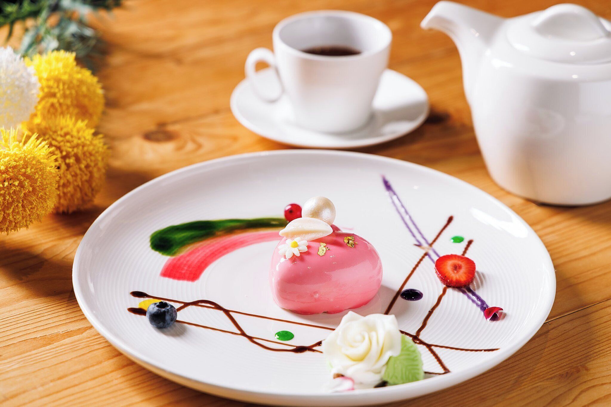 Un piattino con un dessert rosa, una tazza di caffè bianca e un teiera bianca su un tavolo di legno.