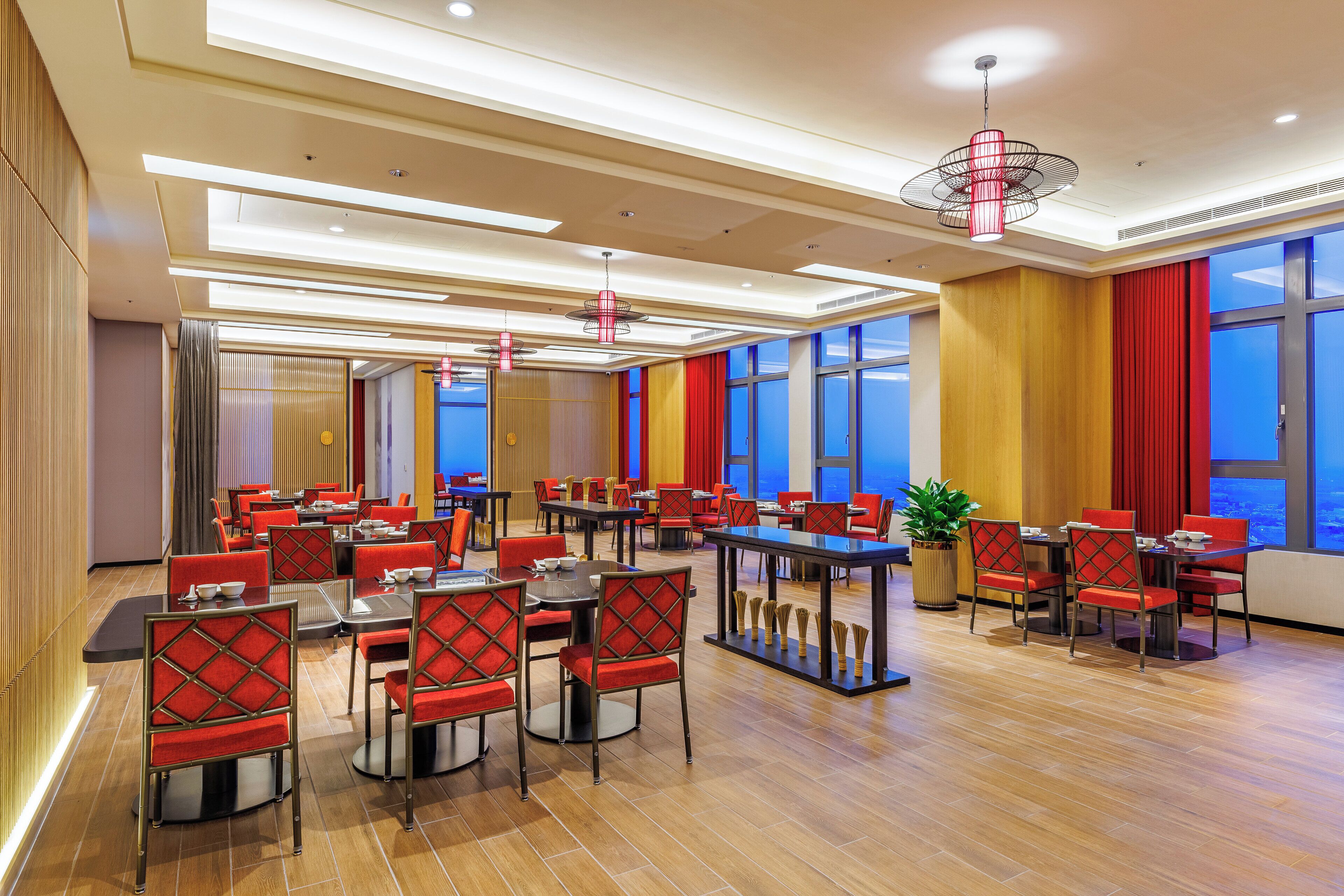 A spacious hotel dining area with multiple tables and chairs, large windows, and modern lighting fixtures.