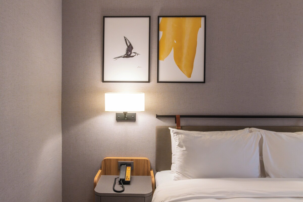 A hotel room with a bed, a nightstand with a lamp, and two framed artworks on the wall.