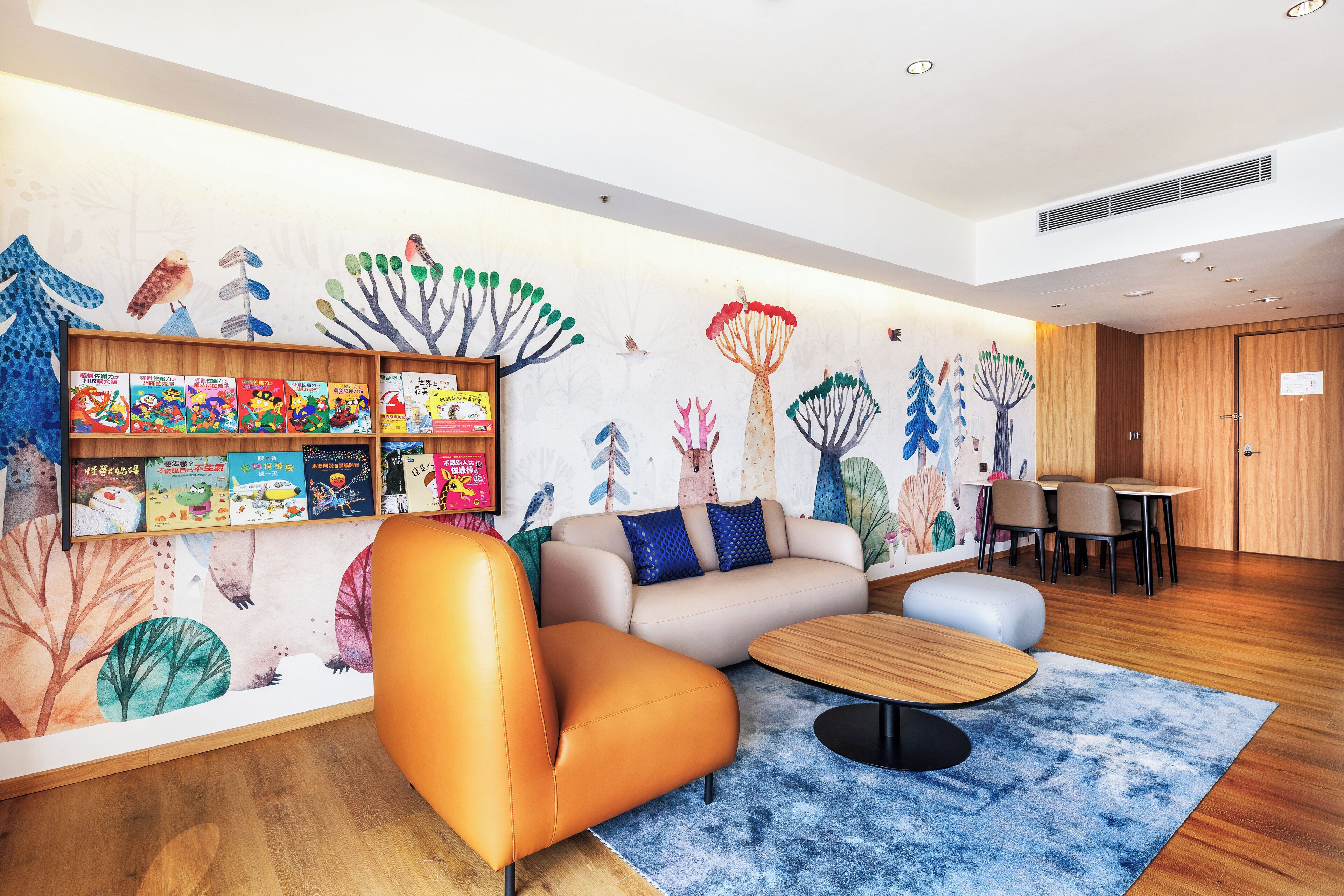A modern hotel lobby with a wooden floor, a sofa, a coffee table, and a bookshelf filled with children's books.