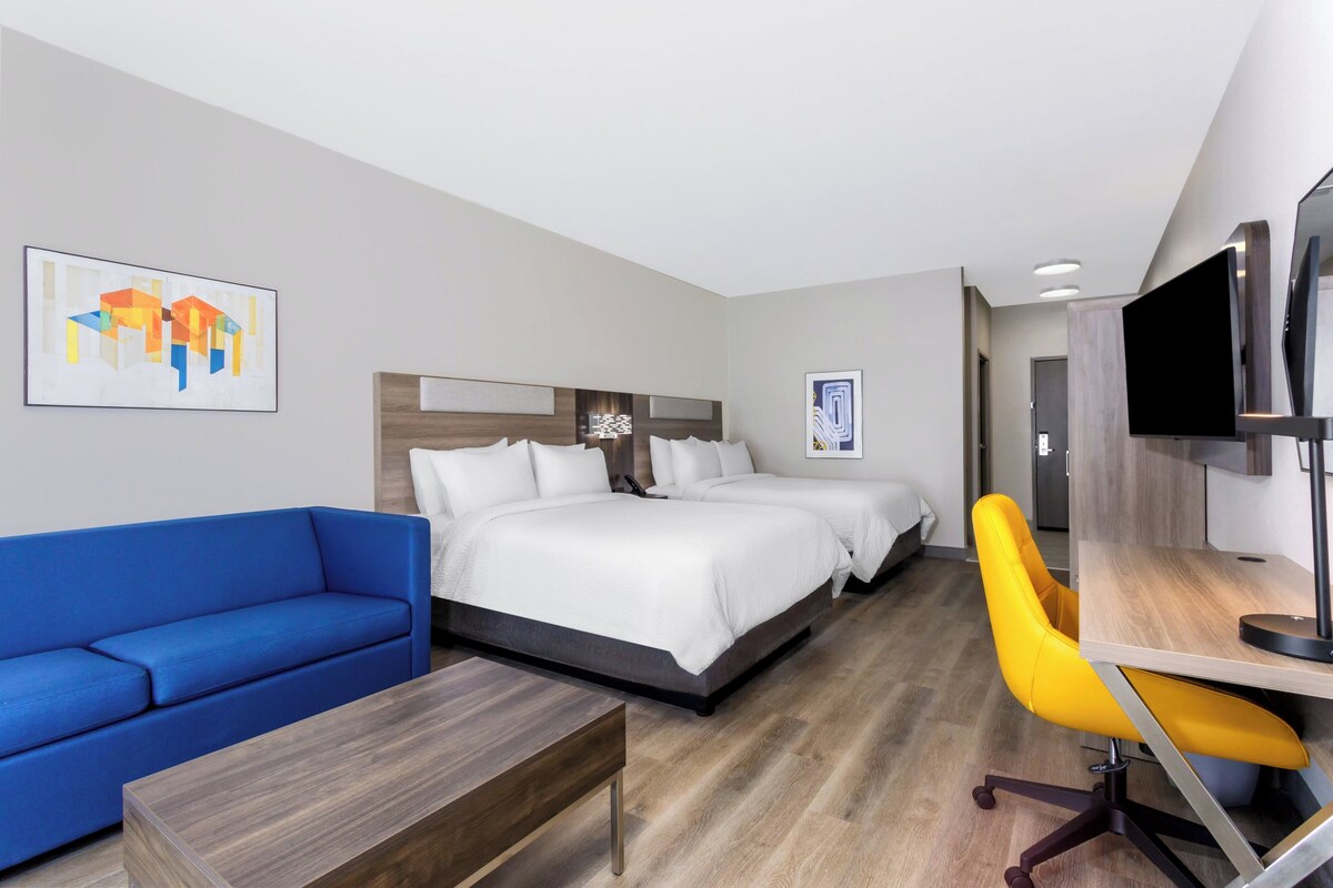 A modern hotel room with a large bed, a blue sofa, a yellow chair, a desk, and a TV.