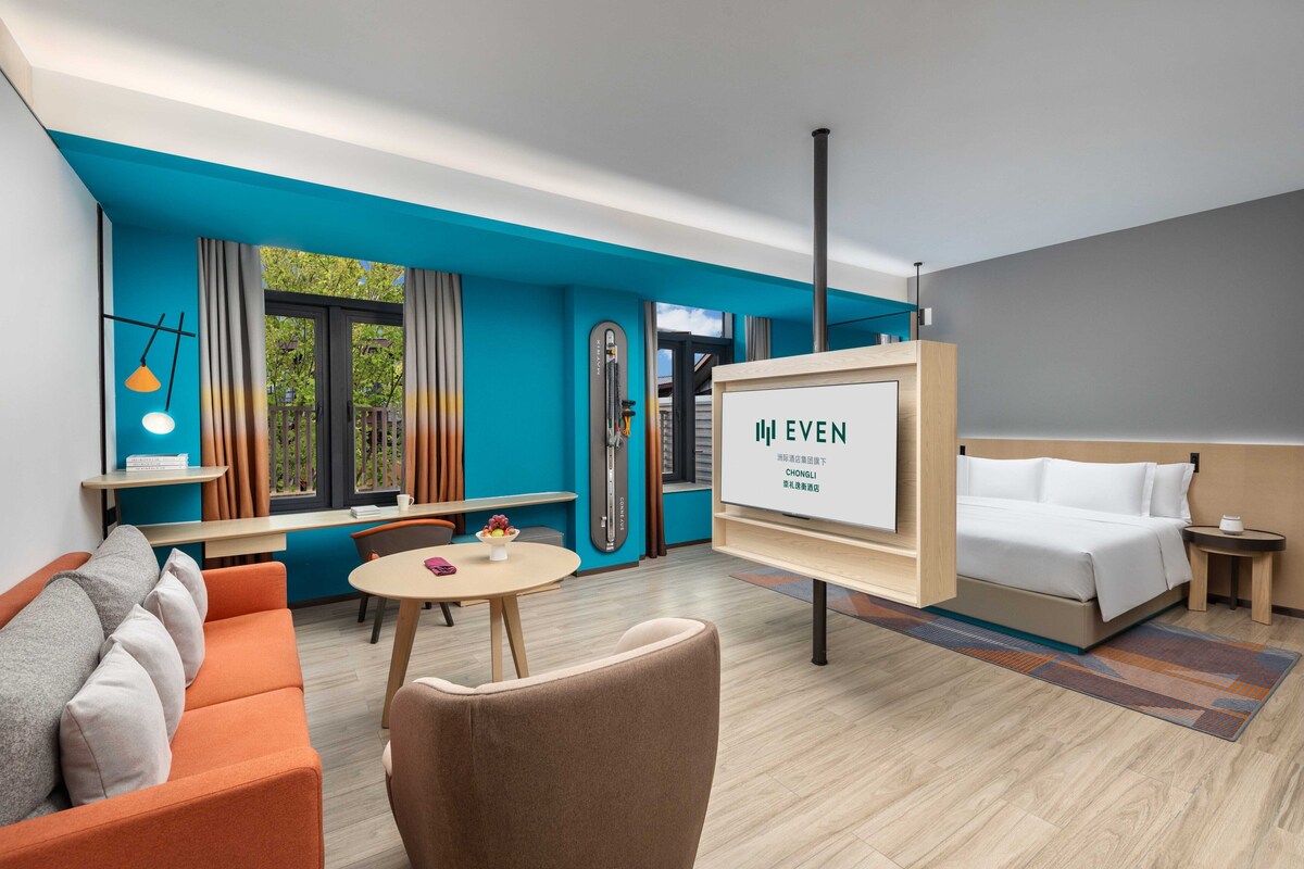 Een moderne hotelkamer met een blauwe accentmuur, een bed, een slaapbank, een eettafel en een televisiescherm waarop de naam van het hotel staat.