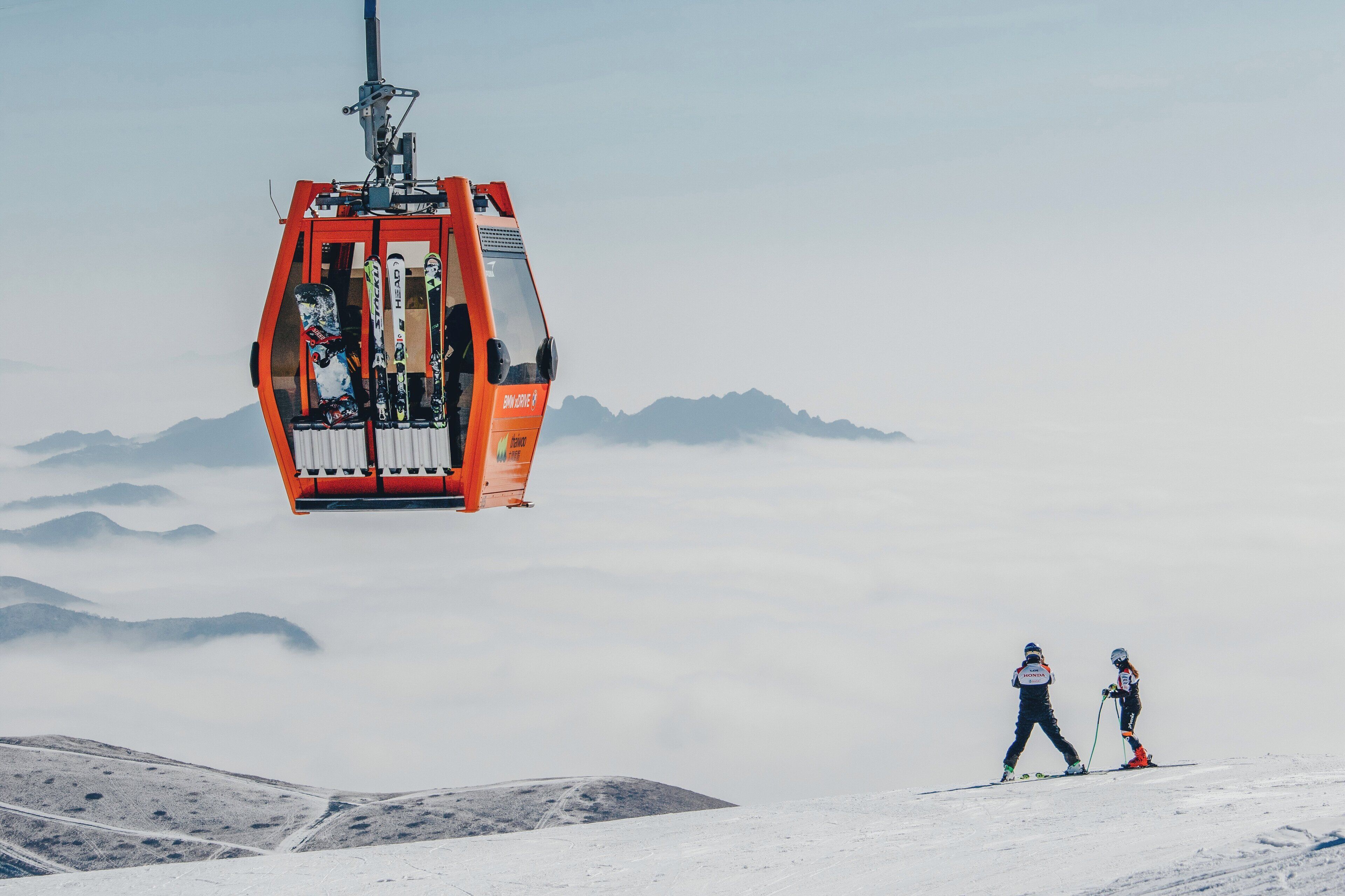 A ski lift with a cabin carrying skiers, set against a backdrop of snow-covered mountains and clouds.