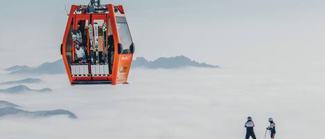 A ski lift with a cabin carrying skiers, set against a backdrop of snow-covered mountains and clouds.