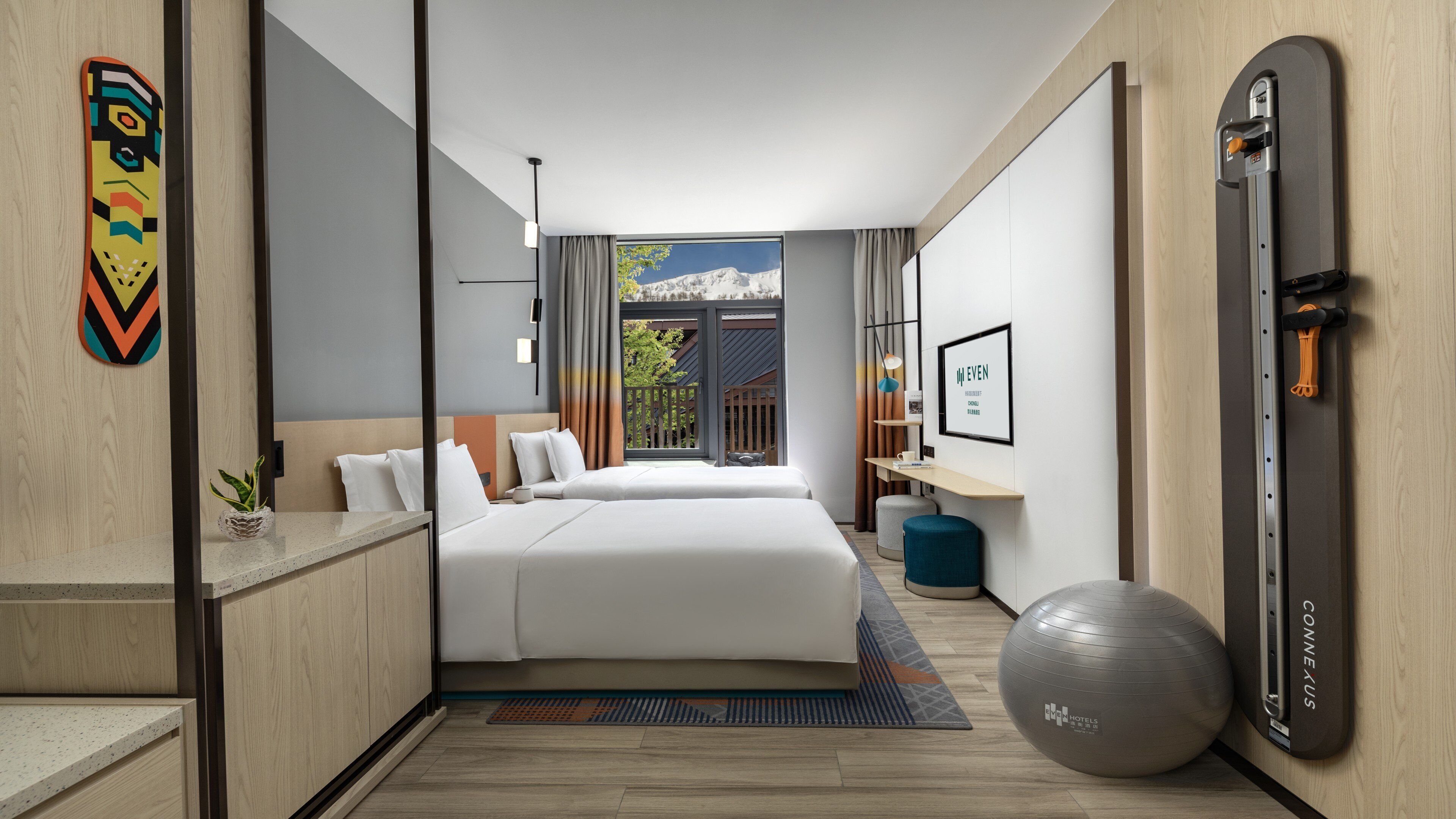A hotel room with a bed, a desk, a chair, a gym ball, a window with a view, and a door with a handle.