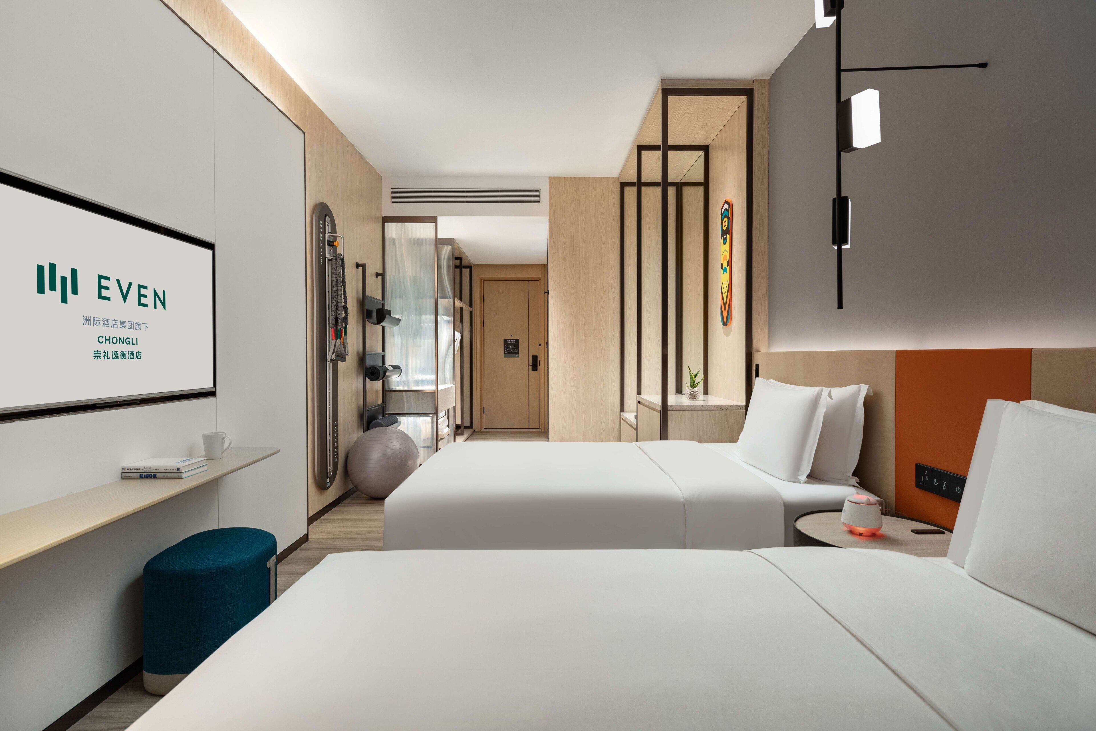 A hotel room with two beds, a desk, a TV, and a wall-mounted exercise ball.