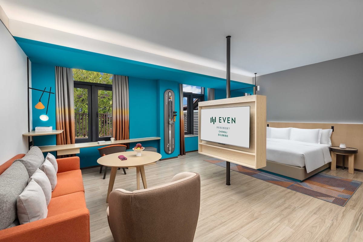 Une chambre d’hôtel moderne avec un mur d’accent bleu, un lit, un canapé, une table à manger et un écran de télévision affichant le nom de l’hôtel.