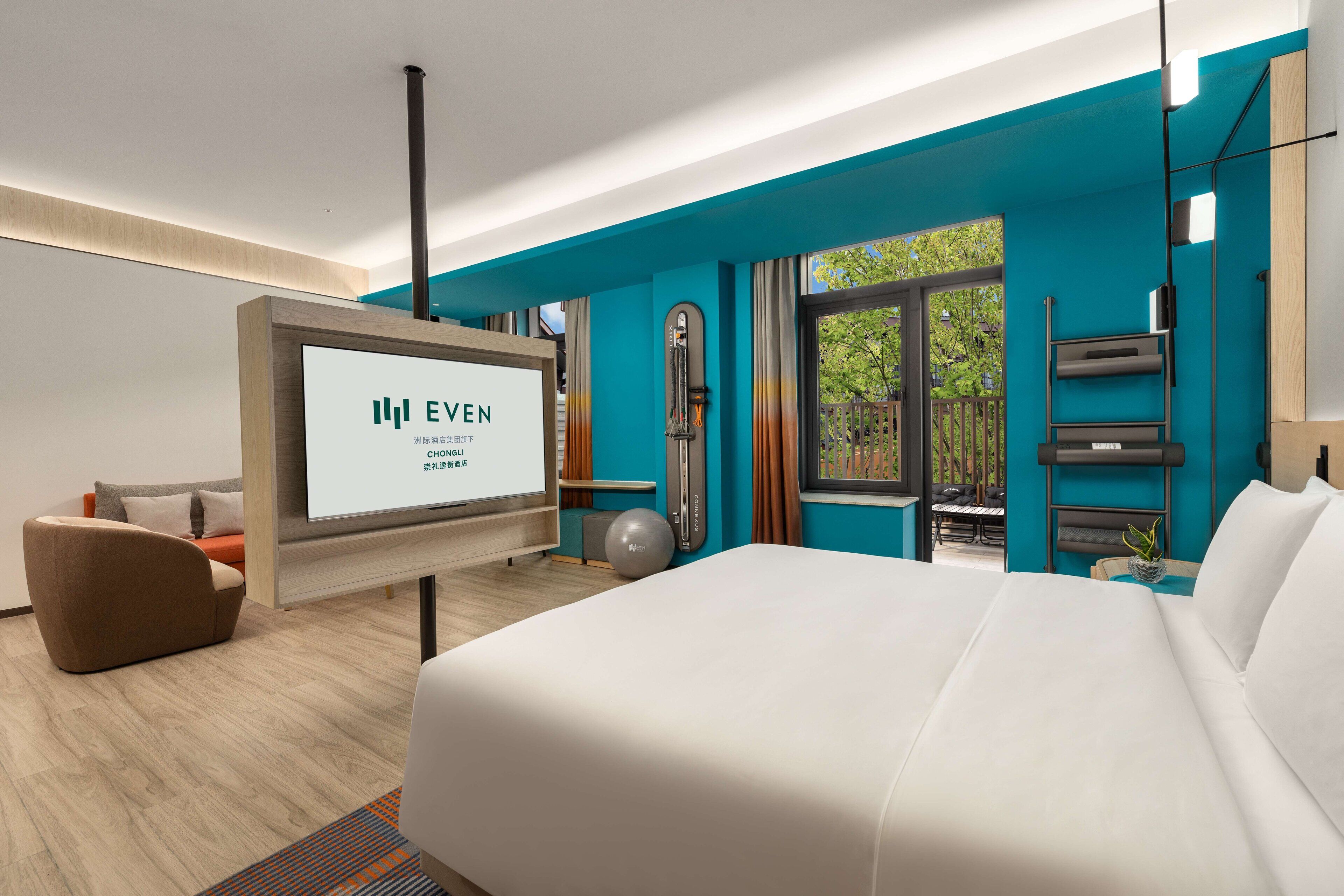 Habitación de hotel moderna con una cama grande, una pantalla de televisión mostrando el nombre del hotel, una pelota de ejercicio y vistas al exterior.