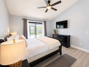 Une chambre à coucher comprenant un lit, un ventilateur de plafond, une télévision, une commode et une fenêtre avec des rideaux.