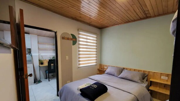 Un dormitorio con techo de madera, cama con ropa de cama gris, mesita de noche y ventana con persianas.