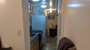 A narrow hallway with a kitchen area at the end, featuring cabinets, a refrigerator, and a tiled floor.