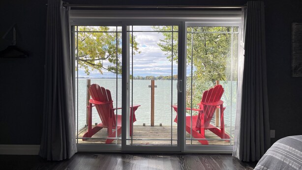 A room with a large sliding glass door leading to a deck with two red Adirondack chairs and a view of a body of water. - Sunset Escape On Westlake. This  B&B offers 2 suites that are rented separately. (Prince Edward)