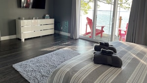 A modern bedroom with a large bed, a white dresser, a flat-screen TV, and a view of the water through large sliding doors. - Sunset Escape On Westlake. This  B&B offers 2 suites that are rented separately. (Prince Edward)