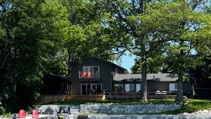A modern two-story house with a deck overlooking a body of water, surrounded by lush greenery. - Sunset Escape On Westlake. This  B&B offers 2 suites that are rented separately. (Prince Edward)