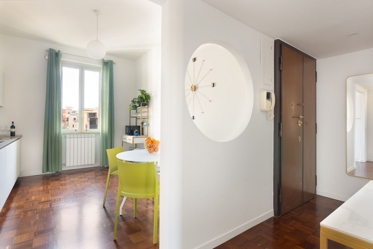 A modern kitchen with a dining area, a large window with curtains, a round wall decoration, and a door with a mirror.