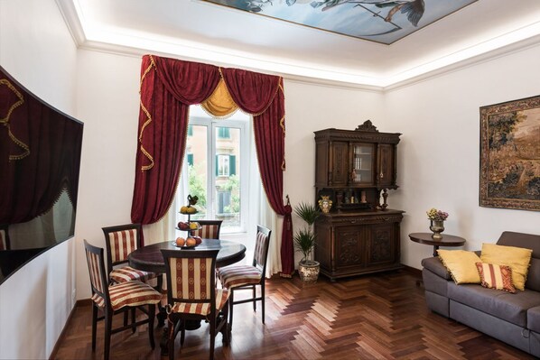 A dining area with a round table, striped chairs, and a view of the outdoors through large windows with red curtains. - Idyllia | Classy apartment in Vatican area (Roma)