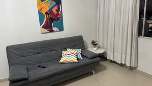 A modern living room with a grey sofa, a colorful cushion, a remote control, a small white table with a vase of flowers, and a large abstract painting on the wall.