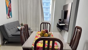 A dining area with a table set for four, surrounded by wooden chairs. A fruit bowl is on the table, and a sofa is in the background.