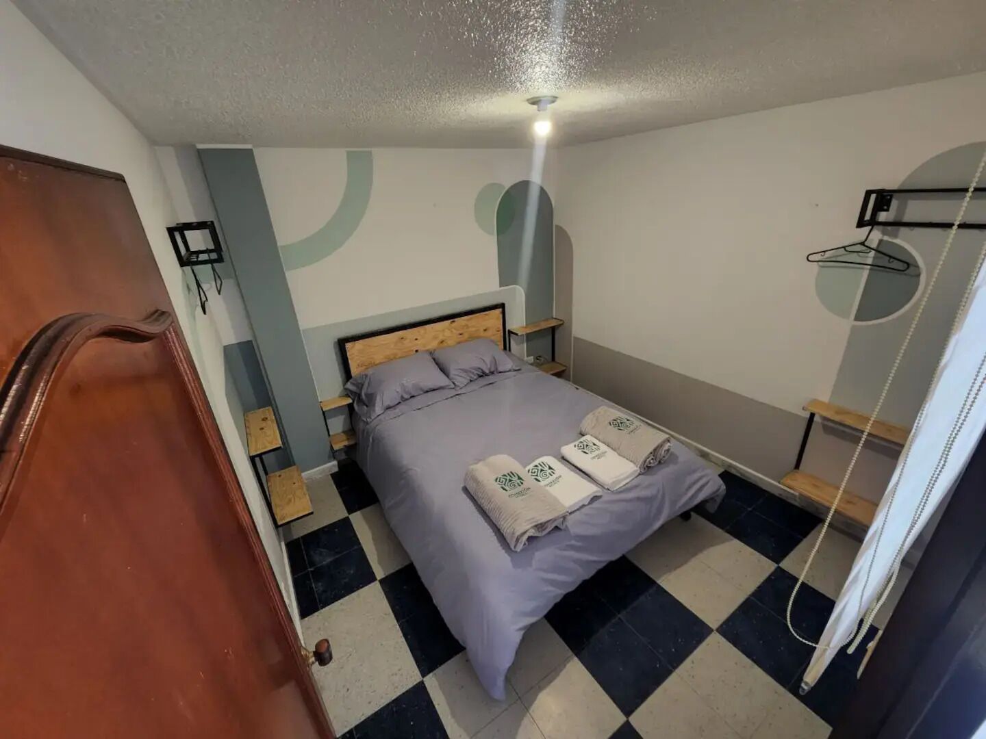 Une petite chambre à coucher, bien rangée, avec un sol à carreaux, un lit avec des oreillers, une table de chevet et un porte-manteau suspendu.