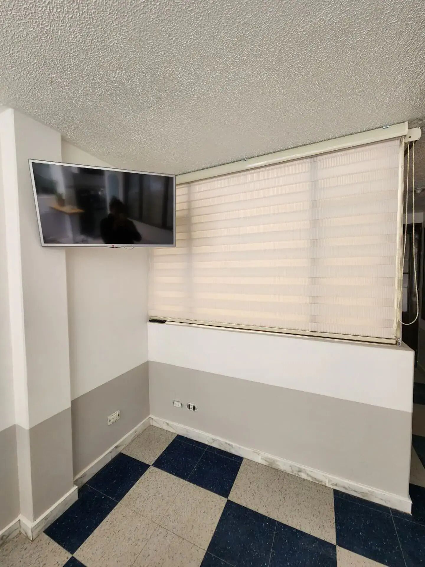 Ein Zimmer mit einem an der Wand befestigten Flachbildfernseher, einem Fenster mit Jalousien, einem Schachbrettmuster Boden und einer niedrigen, weißen und grauen Wand.