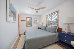 Een slaapkamer met een bed, nachtkastje, lamp, plafondventilator en een raam met jaloezieën.