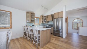 A modern kitchen with wooden cabinets, a central island with chairs, and a large refrigerator. - 4670 - Salted Skyline - Dog-Friendly Coastal Escape Near the Beach (Southern Shores)
