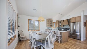 A modern kitchen with wooden flooring, white chairs, a central island, and stainless steel appliances. - 4670 - Salted Skyline - Dog-Friendly Coastal Escape Near the Beach (Southern Shores)