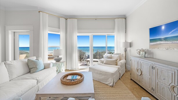 A spacious living room with a large window offering a view of the ocean, a white sofa, a coffee table with a book and a candle, and a white cabinet. - "Beach DNA" at The Legacy 203 in Seagrove Beach | My Beach Getaways (Santa Rosa Beach)