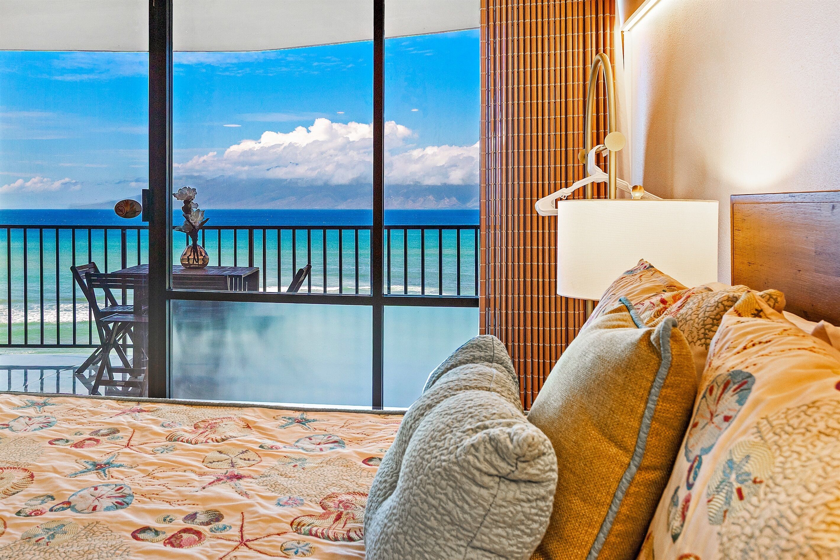 A hotel room with a large window offering a view of the ocean, a bed with patterned bedding, and a small table with chairs outside.