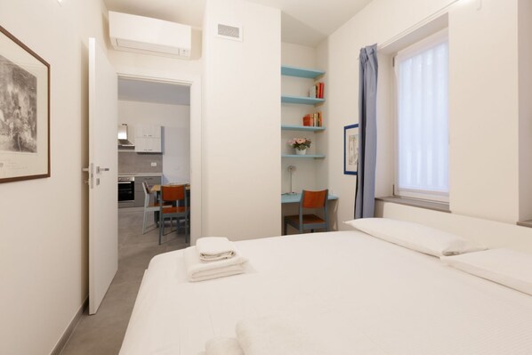 Habitación de hotel con cama, escritorio, silla, kitchenette pequeño y ventana con cortinas.