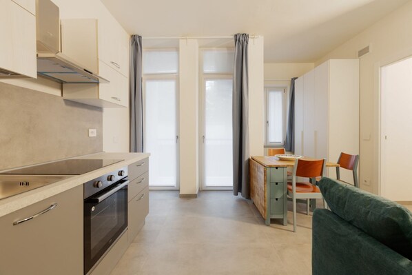 Een kleine keuken met een fornuis, oven en kasten, een eethoek met een tafel en stoelen, en een groene bank.