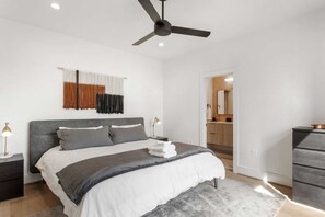 A modern bedroom with a bed, bedside tables, a ceiling fan, and a bathroom visible through an open door.