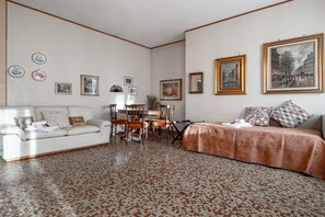 Une pièce avec un sol à motifs, un canapé blanc, un lit recouvert d’une couvre-lit à motifs, une table à manger avec des chaises et divers tableaux encadrés accrochés aux murs.
