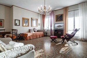 A hotel room with a bed, a sofa, a desk, a chair, a TV, and a chandelier.