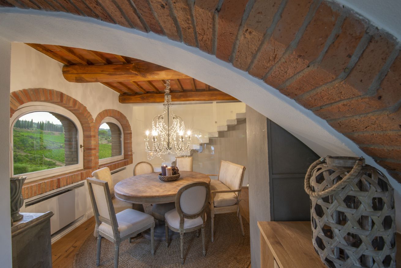 A dining area with a round table, chairs, and a chandelier. There are arched windows and a brick wall.