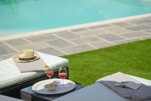 Escena junto a la piscina con una silla de reposo, un sombrero, una bandeja con bebidas y aperitivos, y un libro.