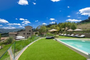 A poolside area with a clear view of a stone building, a grassy lawn, and a row of sun loungers.