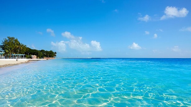 A beach with clear turquoise water, a sandy shore, and a few structures on the left.