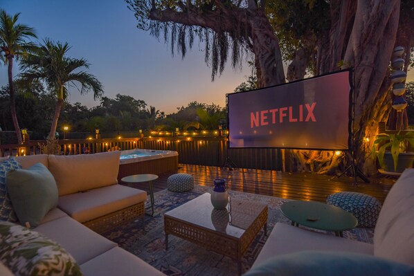 A cozy outdoor seating area with a large screen displaying "NETFLIX" and a hot tub in the background.