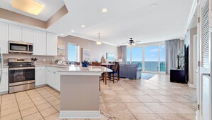 A modern kitchen with white cabinets, stainless steel appliances, and a large island. There is a dining area with a gray sofa and a view of the ocean. - BEACHFRONT, ZERO ENTRY POOL, PRIVATE BALCONY! (Gulf Shores)