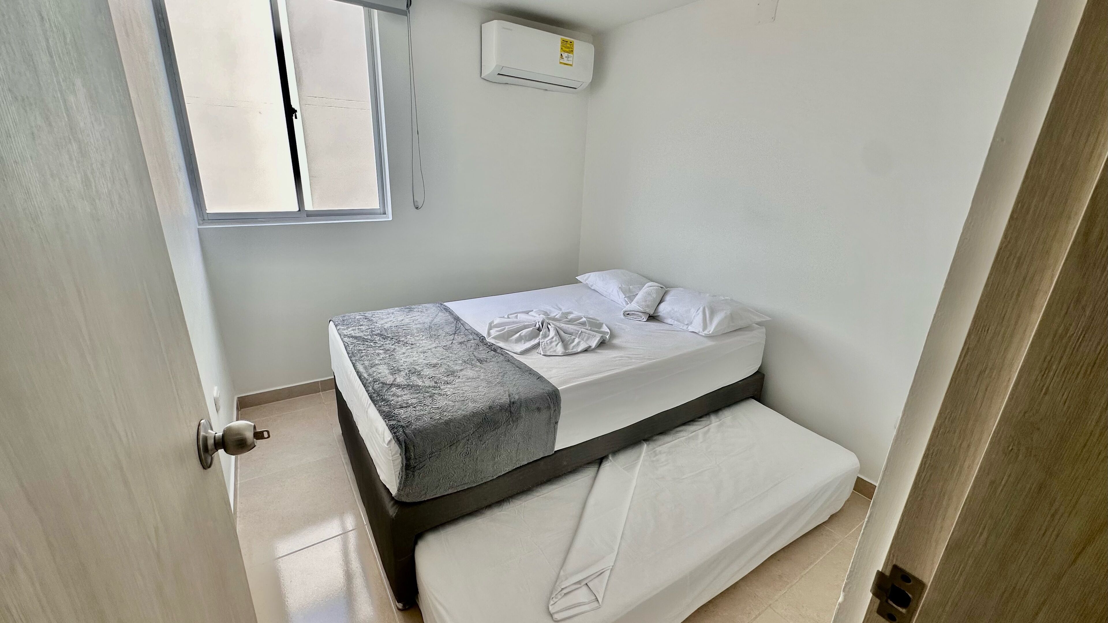 A small, clean room with a single bed, a window, and an air conditioning unit.