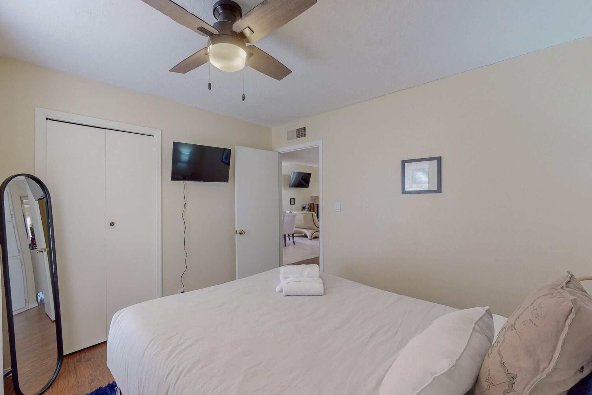 A bedroom with a bed, a ceiling fan, a TV mounted on the wall, and a door leading to another room.