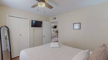A bedroom with a bed, a ceiling fan, a TV mounted on the wall, and a door leading to another room.